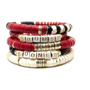 May include: Stack of red, white, and black beaded bracelets with gold accents. The bracelets spell out "BOOMER" and "SOONER" in white letters with gold trim.