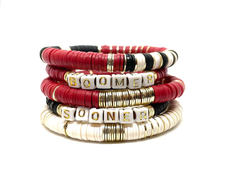 May include: Stack of red, white, and black beaded bracelets with gold accents. The bracelets spell out "BOOMER" and "SOONER" in white letters with gold trim.