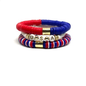May include: Three stacked bracelets with red, white and blue beads. The middle bracelet has gold beads and white beads with the letters "USA" spelled out.