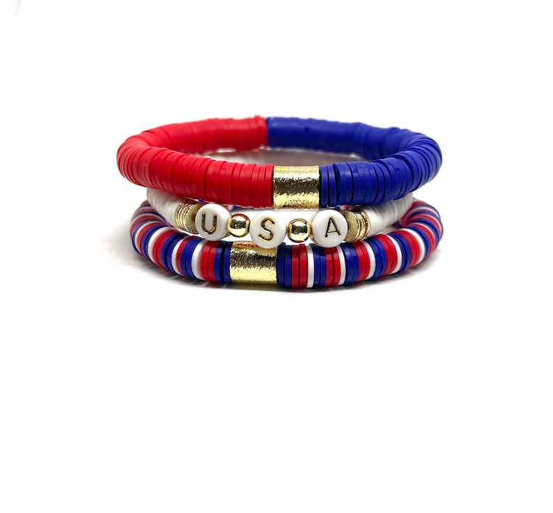 May include: Three stacked bracelets with red, white and blue beads. The middle bracelet has gold beads and white beads with the letters "USA" spelled out.