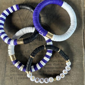 May include: Three colorful bracelets made of round beads. The bracelets are stacked on top of each other. The top bracelet is purple and white. The middle bracelet is black and white. The bottom bracelet is purple, black, and white. The bottom bracelet has gold beads and the letters "HORNED FROG" spelled out in gold letters.