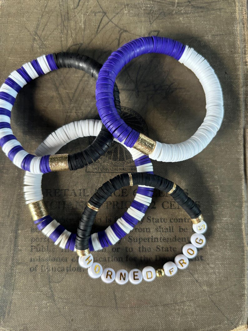 May include: Three colorful bracelets made of round beads. The bracelets are stacked on top of each other. The top bracelet is purple and white. The middle bracelet is black and white. The bottom bracelet is purple, black, and white. The bottom bracelet has gold beads and the letters "HORNED FROG" spelled out in gold letters.