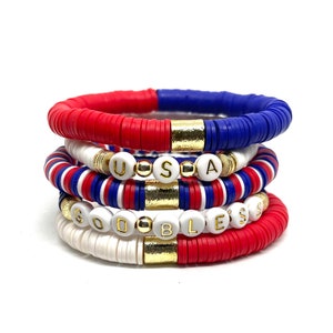 May include: A stack of five bracelets with red, white and blue beads. The top bracelet has gold beads and the bottom bracelet spells out "GOD BLESS" in white beads.