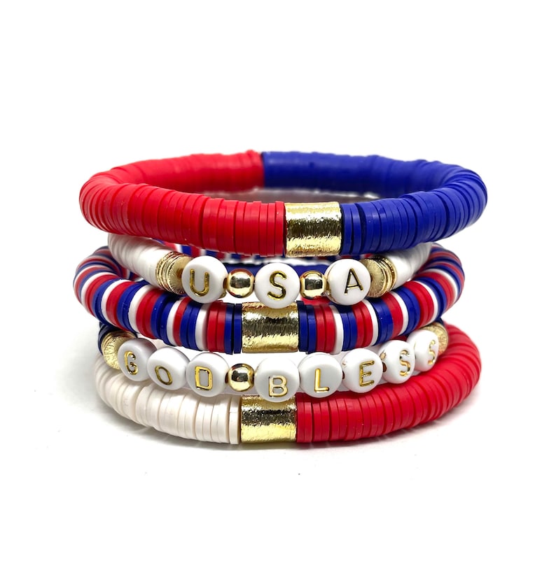 May include: A stack of five bracelets with red, white and blue beads. The top bracelet has gold beads and the bottom bracelet spells out "GOD BLESS" in white beads.