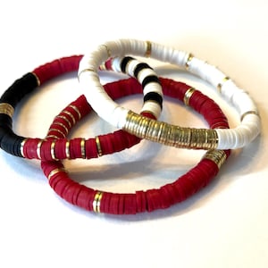 May include: Three bracelets made of colorful ceramic beads with gold accents. The bracelets are red, white, and black.