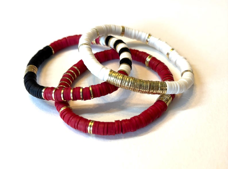 May include: Three bracelets made of colorful ceramic beads with gold accents. The bracelets are red, white, and black.