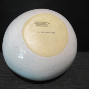 May include: A speckled white ceramic bowl with a light yellow interior base. The bottom of the bowl has text that reads "MADE IN PORTUGAL", "HANDWASH ONLY", and "NOT MICROWAVE SAFE" with the brand name "by ANTHROPOLOGIE".