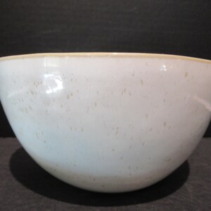 May include: A white ceramic bowl with a speckled pattern and a light tan rim. The bowl has a rounded shape and is likely used for serving food or as a decorative piece. The bowl is sitting on a dark surface.