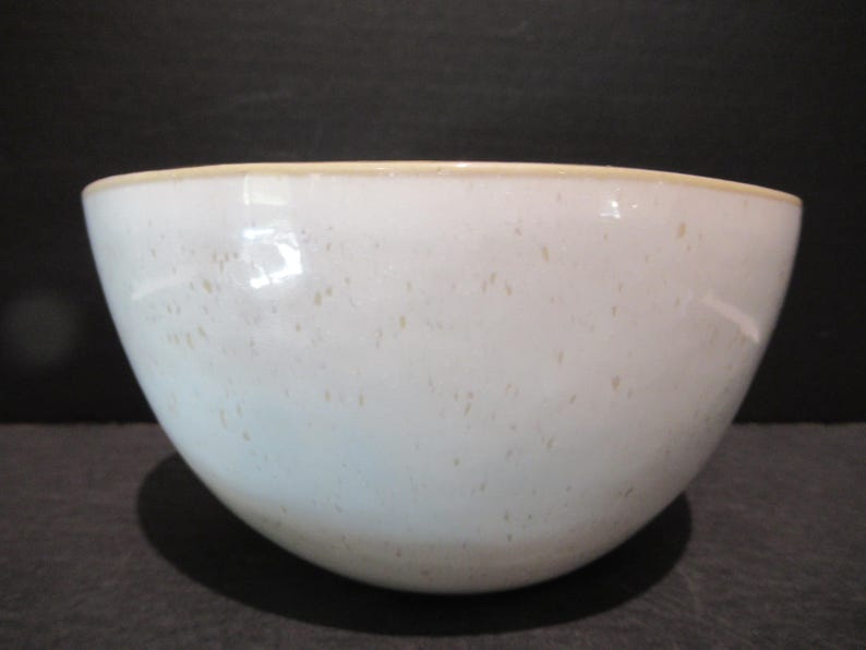 May include: A white ceramic bowl with a speckled pattern and a light tan rim. The bowl has a rounded shape and is likely used for serving food or as a decorative piece. The bowl is sitting on a dark surface.