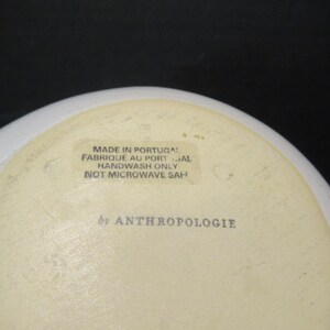 May include: A white ceramic bowl with a cream-coloured interior. The base of the bowl features text that reads "MADE IN PORTUGAL", "HANDWASH ONLY", and "by ANTHROPOLOGIE". The bowl is not microwave safe.