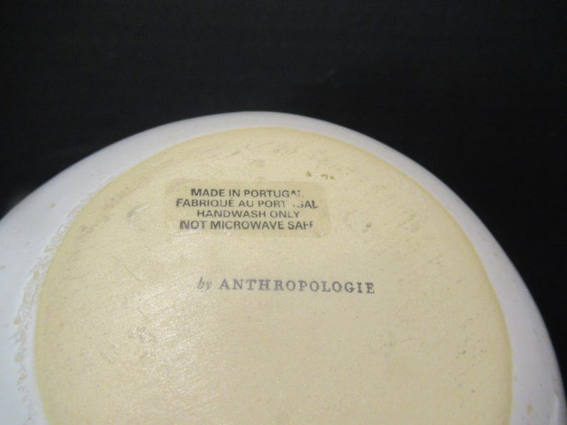 May include: A white ceramic bowl with a cream-coloured interior. The base of the bowl features text that reads "MADE IN PORTUGAL", "HANDWASH ONLY", and "by ANTHROPOLOGIE". The bowl is not microwave safe.