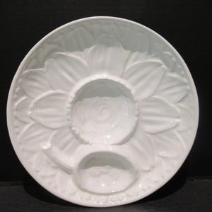 May include: White ceramic serving platter with a sunflower design. The platter features a central dip bowl and two additional compartments. The sunflower petals are raised, adding texture to the serving dish. Ideal for serving appetizers or dips.