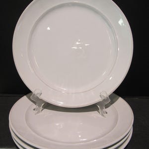 May include: A stack of four white ceramic plates with a wide rim. One plate is elevated on a clear stand, showcasing the clean, minimalist design. The plates are suitable for serving meals.