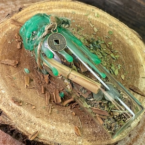 May include: A clear glass bottle filled with dried herbs, bark, and a rolled-up scroll. The bottle is sealed with green wax and tied with twine, and a small coin charm hangs from the twine. The bottle rests on a wooden surface.