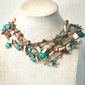 May include: A multi-strand necklace displayed on a mannequin. The necklace features turquoise, pink, white, and brown beads, along with small turtle charms. The strands are connected by a brown cord, creating a layered effect.