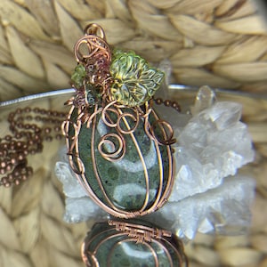 May include: A copper wire wrapped pendant featuring a green and white moss agate stone with green glass leaves. The pendant is on a copper chain.