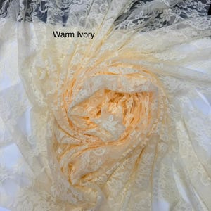 May include: A sheer, warm ivory lace fabric featuring a delicate floral pattern. The material is gathered into a central spiral, showcasing the intricate, semi-transparent mesh construction and the soft, light-toned aesthetic of the textile.