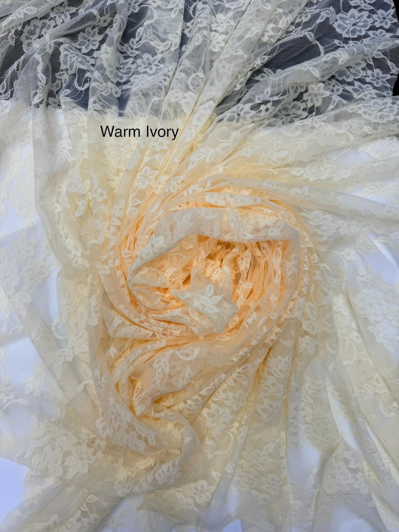 May include: A sheer, warm ivory lace fabric featuring a delicate floral pattern. The material is gathered into a central spiral, showcasing the intricate, semi-transparent mesh construction and the soft, light-toned aesthetic of the textile.