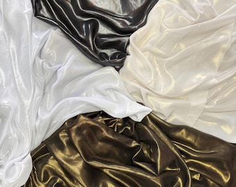 May include: An overhead shot shows several swatches of shiny fabric in various colours. The colours include black, white, gold, and a bronze-like shade. The fabric is draped and folded, creating a textured appearance.