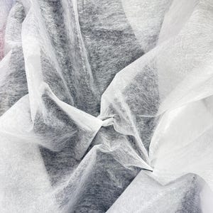 May include: Close-up of white, translucent fabric with a soft, flowing texture. The fabric is layered and draped, creating shadows and highlights. The background is a darker gray, providing contrast to the white material. The image suggests a textile or craft material.