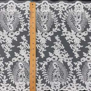 May include: White lace fabric with a repeating pattern of the Virgin Mary and floral motifs. The lace is measured with a wooden ruler, showcasing a detailed, openwork design. The fabric is suitable for craft and sewing projects. The ruler shows approximately 48 cm of fabric.