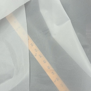 May include: A close-up shot of white sheer fabric with a wooden ruler measuring from 12 to 22 inches. The fabric is folded and draped, creating soft shadows and textures. The ruler is angled across the fabric.