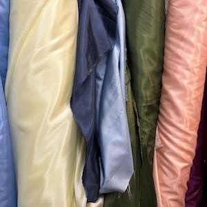 May include: A close-up of several rolls of fabric in various colors. The colors include light blue, pale yellow, navy blue, olive green, peach, and burgundy. The fabric appears to be a smooth, shiny material, possibly satin or silk.