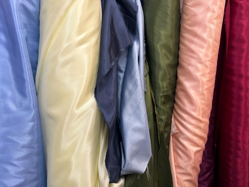 Puede incluir: Primer plano de varios rollos de tela en varios colores. Los colores incluyen azul claro, amarillo p&aacute;lido, azul marino, verde oliva, melocot&oacute;n y burdeos. La tela parece ser un material liso y brillante, posiblemente sat&eacute;n o seda.