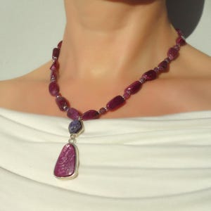 Op de afbeelding: Een roze en paarse edelsteen ketting met een hanger met een grotere roze edelsteen. De ketting is gemaakt van gefacetteerde kralen en wordt gedragen door een model.