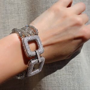 Puede incluir: Un brazalete plateado con un cierre cuadrado engastado con piedras transparentes. El brazalete está hecho de múltiples hilos de cuentas grises.