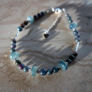 May include: A beaded bracelet featuring a mix of blue, purple, and brown gemstones. The bracelet has a silver chain and a small heart charm. The gemstones vary in size and shape, creating a unique and eye-catching design.