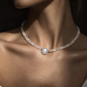 May include: A beaded necklace featuring small, translucent, iridescent round beads and a central circular pendant with a faceted, shimmering white stone set in a thin silver-toned metal frame, resting against the base of the neck.