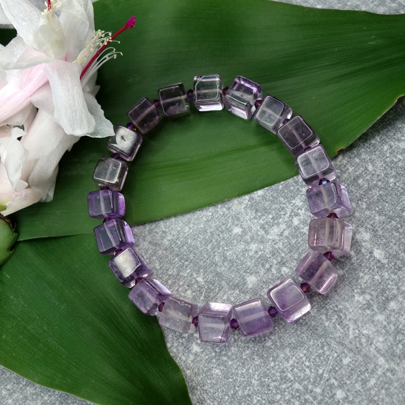 May include: A purple amethyst gemstone bracelet with square-shaped beads. The bracelet is on a green leaf.