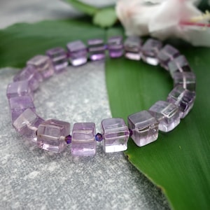 May include: A purple amethyst gemstone bracelet with square-shaped beads. The bracelet is made with faceted beads and is strung on a clear elastic cord.