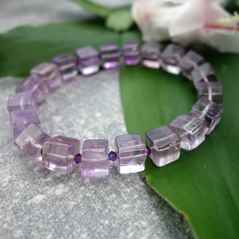 May include: A purple amethyst gemstone bracelet with square-shaped beads. The bracelet is made with faceted beads and is strung on a clear elastic cord.