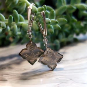 Peut inclure: Une paire de boucles d'oreilles en argent avec un pendentif carré de couleur gris foncé, facetté, orné d'un motif floral. Chaque pendentif est attaché à une chaîne en argent avec une perle de cristal transparente.