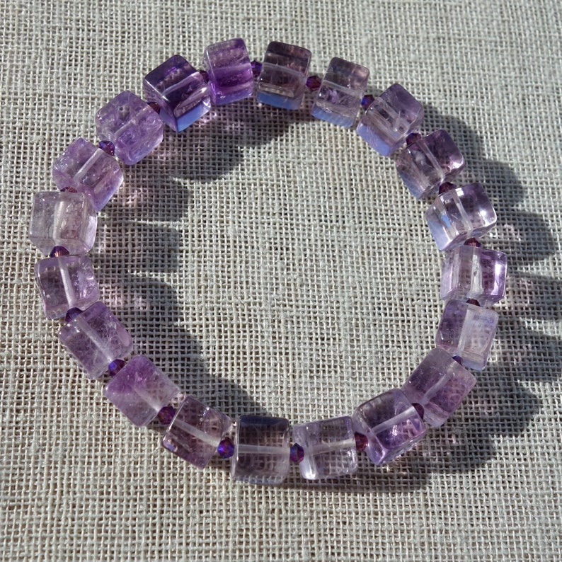 May include: A purple amethyst gemstone bracelet with square-shaped beads. The bracelet is made with small silver beads between each gemstone.