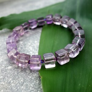 May include: A purple amethyst gemstone bracelet with square-shaped beads. The bracelet is on a green leaf.