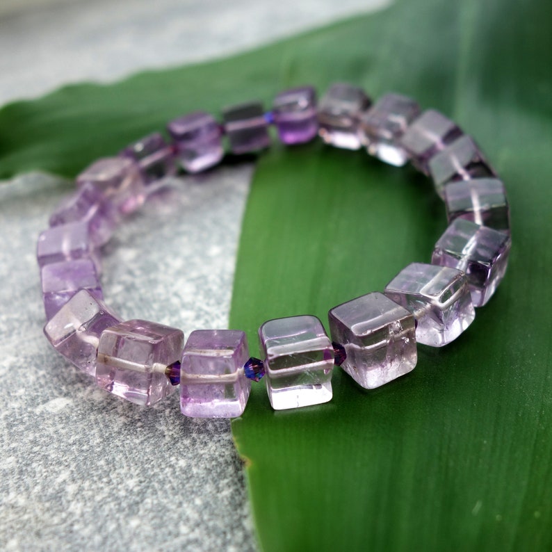 May include: A purple amethyst gemstone bracelet with square-shaped beads. The bracelet is on a green leaf.