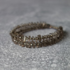 May include: A double-strand bracelet featuring smoky quartz and small metallic beads. The bracelet is a neutral brown color and appears to be strung together with a thin, flexible cord. The beads are rectangular and irregular in shape.