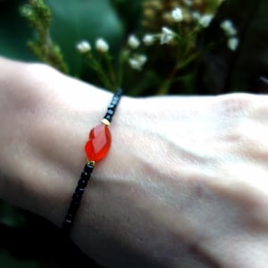 Può includere: Un braccialetto delicato con un unico ciondolo di cornalina sfaccettato a forma di cuore. Il braccialetto è realizzato con perline nere e dettagli dorati.