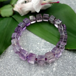 May include: A purple amethyst gemstone bracelet with square-shaped beads. The bracelet is on a green leaf.