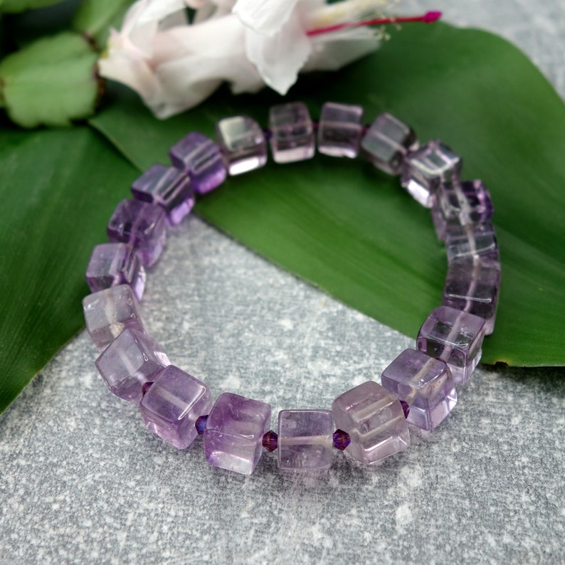 May include: A purple amethyst gemstone bracelet with square-shaped beads. The bracelet is on a green leaf.