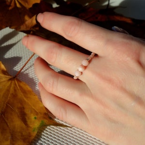 Op de afbeelding: Een delicate ring gemaakt van roze opaal kralen geregen aan een zilveren draad. De ring wordt gedragen op een hand met een lichte huidskleur.