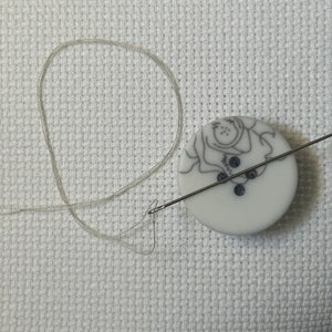 Può includere: Un bottone bianco con un disegno floreale bianco e nero e tre bottoni neri. Un ago e un filo bianco sono infilati nel bottone.