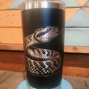 May include: Black stainless steel tumbler with a silver engraved rattlesnake design. The tumbler has a clear lid and a silver rim at the top and bottom. The snake is coiled and has its tongue out.