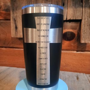 May include: Black stainless steel tumbler with a silver cross design. The cross features a list of album titles including "Blizzard of Ozz" and "Diary of a Madman". The tumbler has a clear lid and sits on a light blue base.