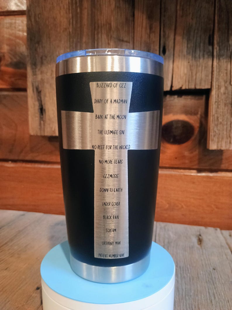 May include: Black stainless steel tumbler with a silver cross design. The cross features a list of album titles including "Blizzard of Ozz" and "Diary of a Madman". The tumbler has a clear lid and sits on a light blue base.