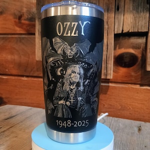 May include: Black stainless steel tumbler featuring the name "OZZY" at the top. The tumbler has an engraved image of a musician and the text "1948-2025" at the bottom. The tumbler is sitting on a light blue and white stand.