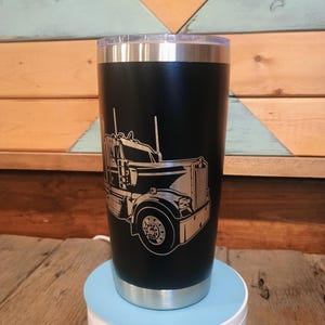 May include: Black insulated tumbler with a silver rim and lid. The tumbler features a detailed etched design of a semi-truck. The tumbler is sitting on a white and light blue rotating stand. The background is a wooden wall.